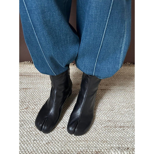 Black tabi-style ankle boots worn with blue jeans on a textured beige woven rug.