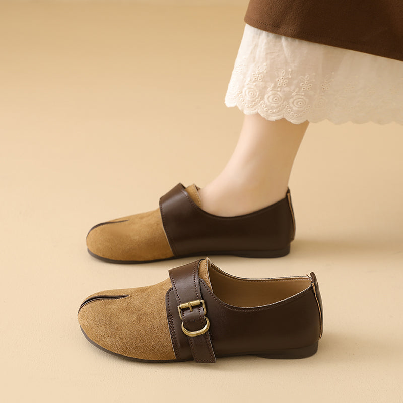 A pair of women's flat shoes in brown and tan suede, shown on a model's feet against a warm beige background. The shoes feature a dark brown strap with a gold-tone buckle across the instep and a tan suede toe section.