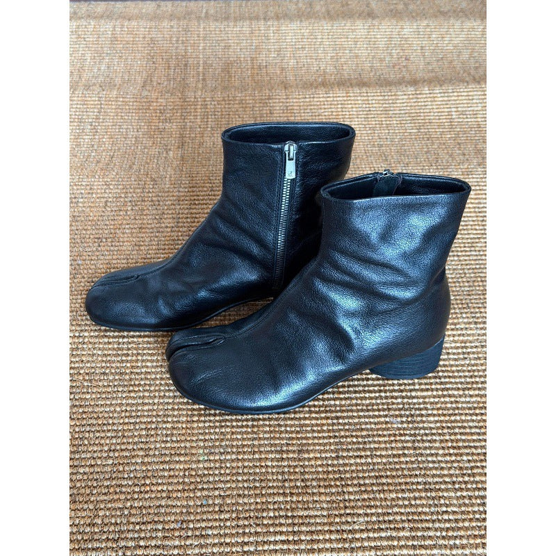 A pair of black leather tabi-style ankle boots with side zippers, displayed on a brown woven mat background.