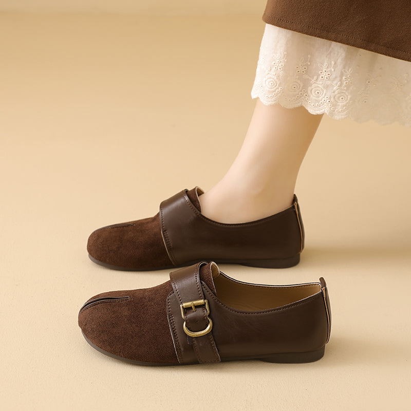 A pair of dark brown suede-and-leather flat shoes with a buckle strap, shown on a model's feet against a warm beige background.