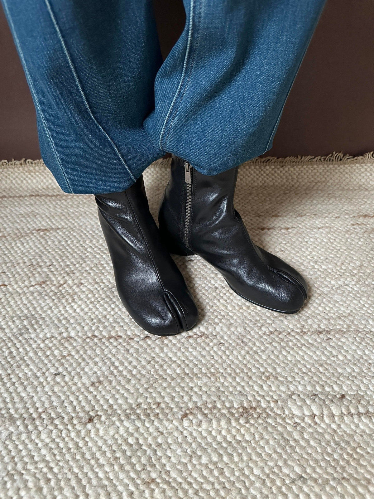 Black leather tab-top ankle boots with side zippers, shown on a person wearing blue jeans standing on a woven beige rug.