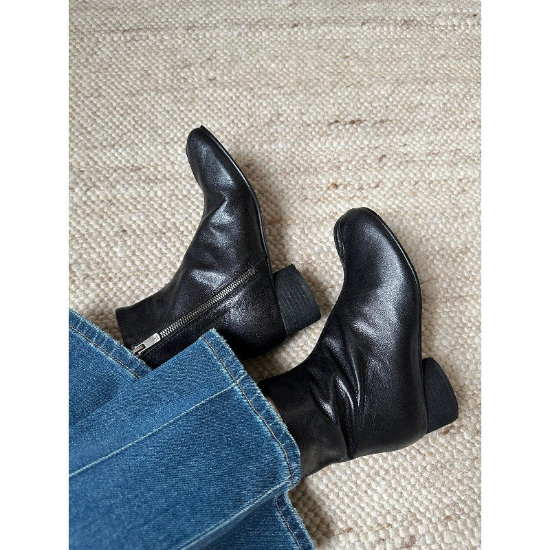 Black leather ankle boots with a side zipper and low block heel, shown on a person's feet wearing blue jeans on a textured beige rug.