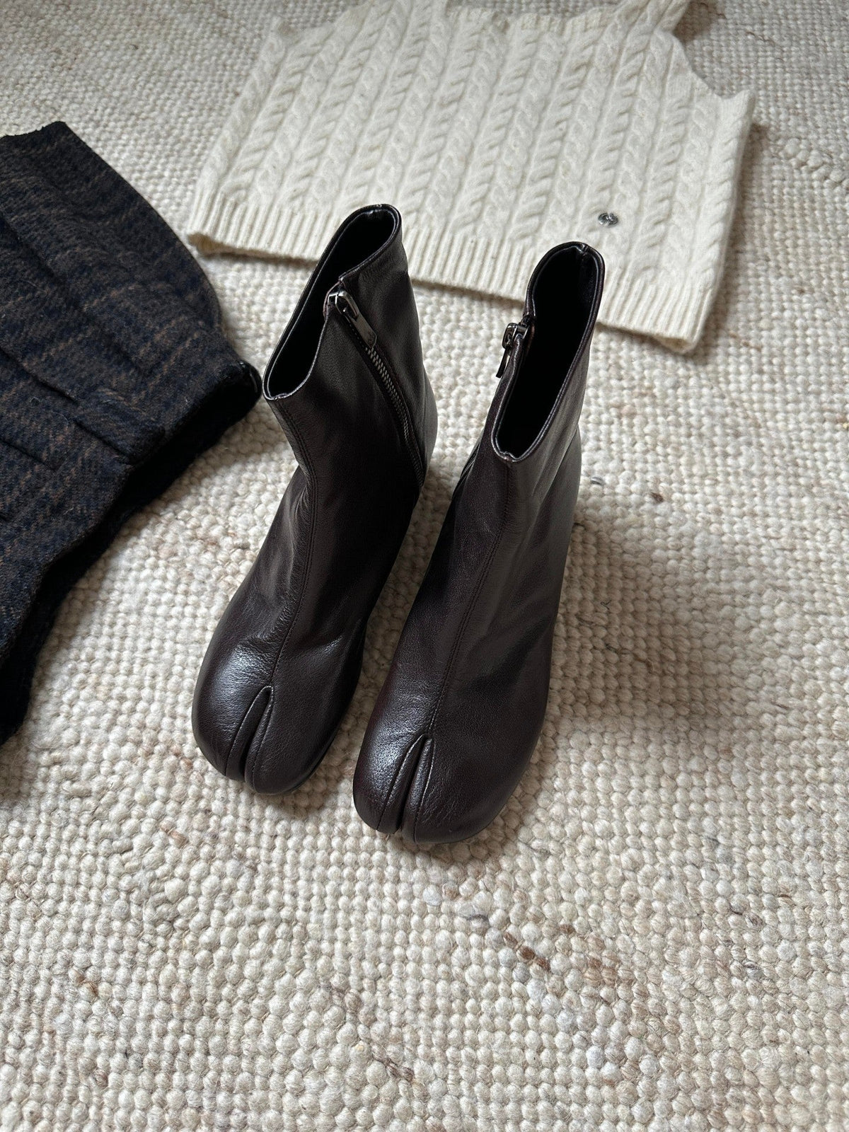 A pair of dark brown leather tab-top ankle boots with side zippers, displayed on a textured beige woven rug alongside a cream cable-knit sweater and a dark knit garment.
