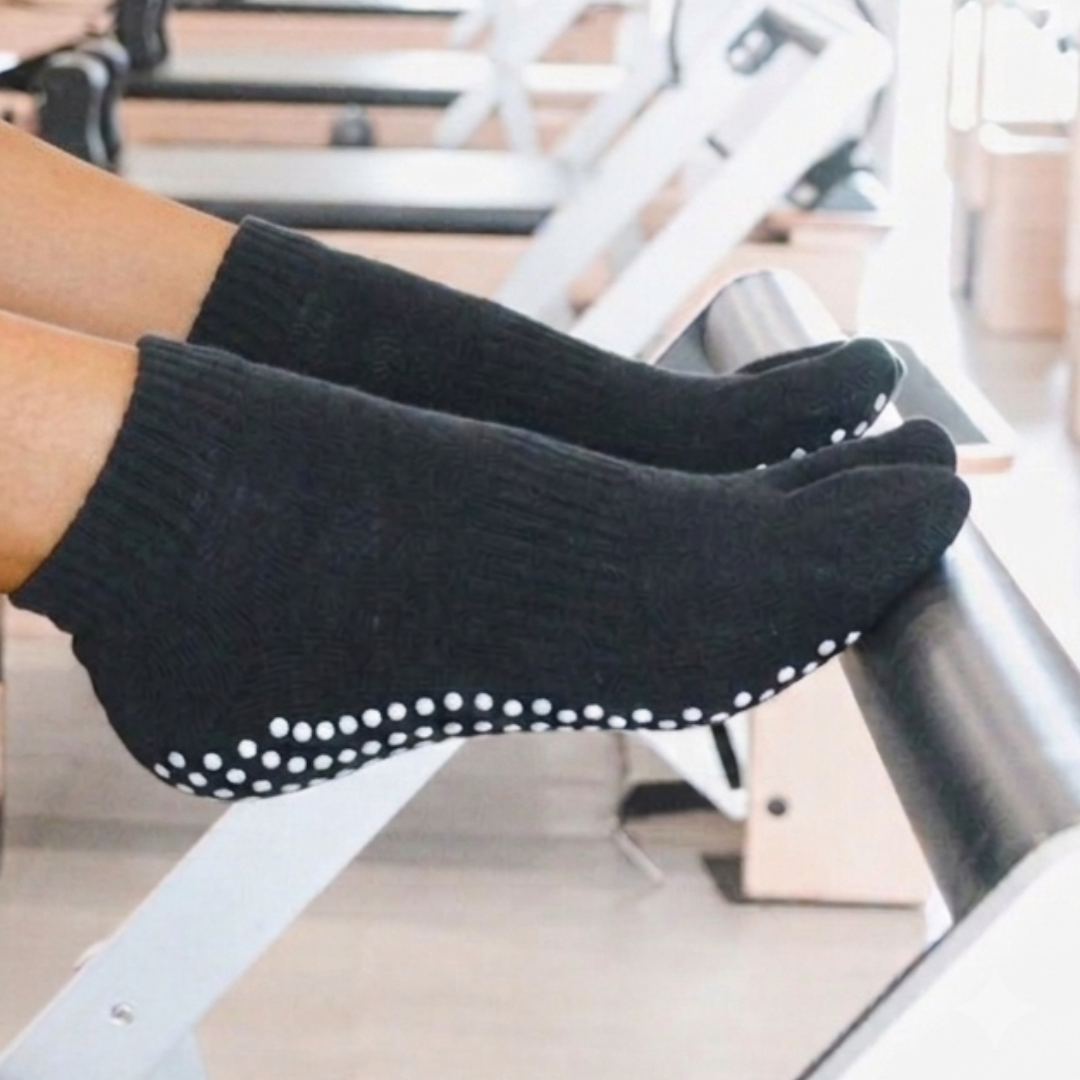 Black grip Tabi V-TOE socks with white dotted non-slip soles worn on feet resting on a Pilates reformer machine in a gym.