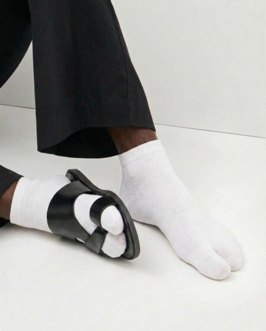 White ankle split-toe tabi socks worn with black slide sandals and black trousers on a clean white floor.
