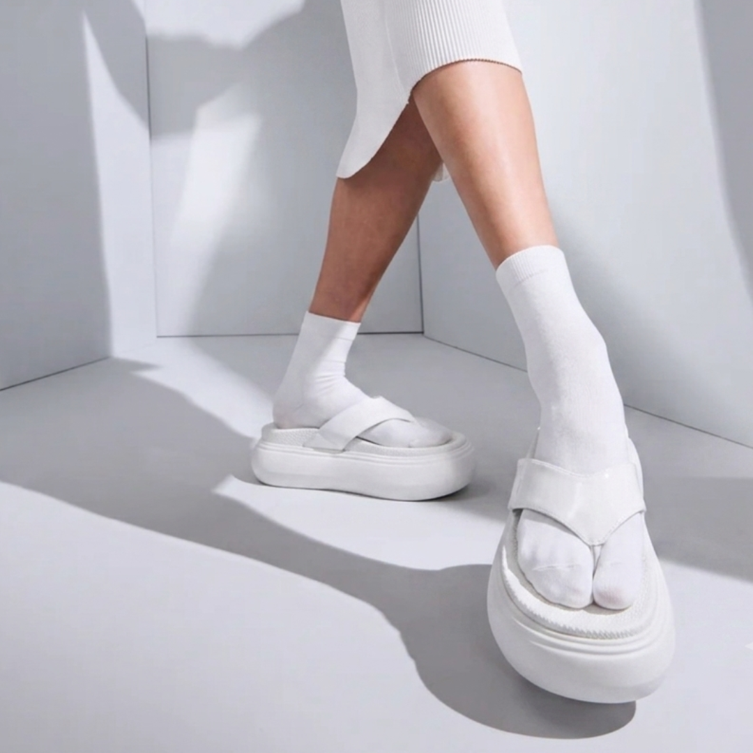 White platform flip flop sandals with wide straps, shown on a model wearing white V-Toe Tabi socks in a minimalist white studio setting.
