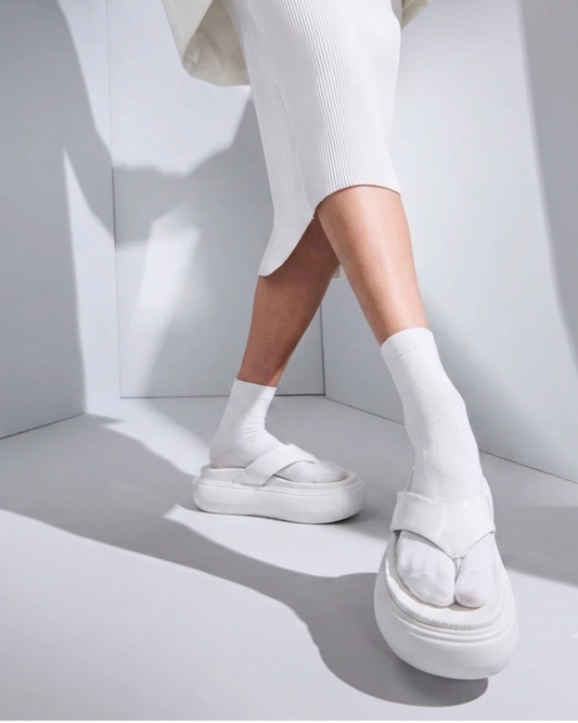 White platform sandals with toe loops and wide straps, shown on a model wearing white V-Toe Tabi socks in a minimalist white studio setting.