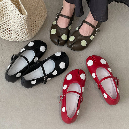 Four tabi-style Mary Jane shoes with large polka dots are arranged on a light neutral surface, with a person's feet wearing one pair in the background. The shoes come in black, dark brown, red, and pinkish red, each with contrasting white dots.