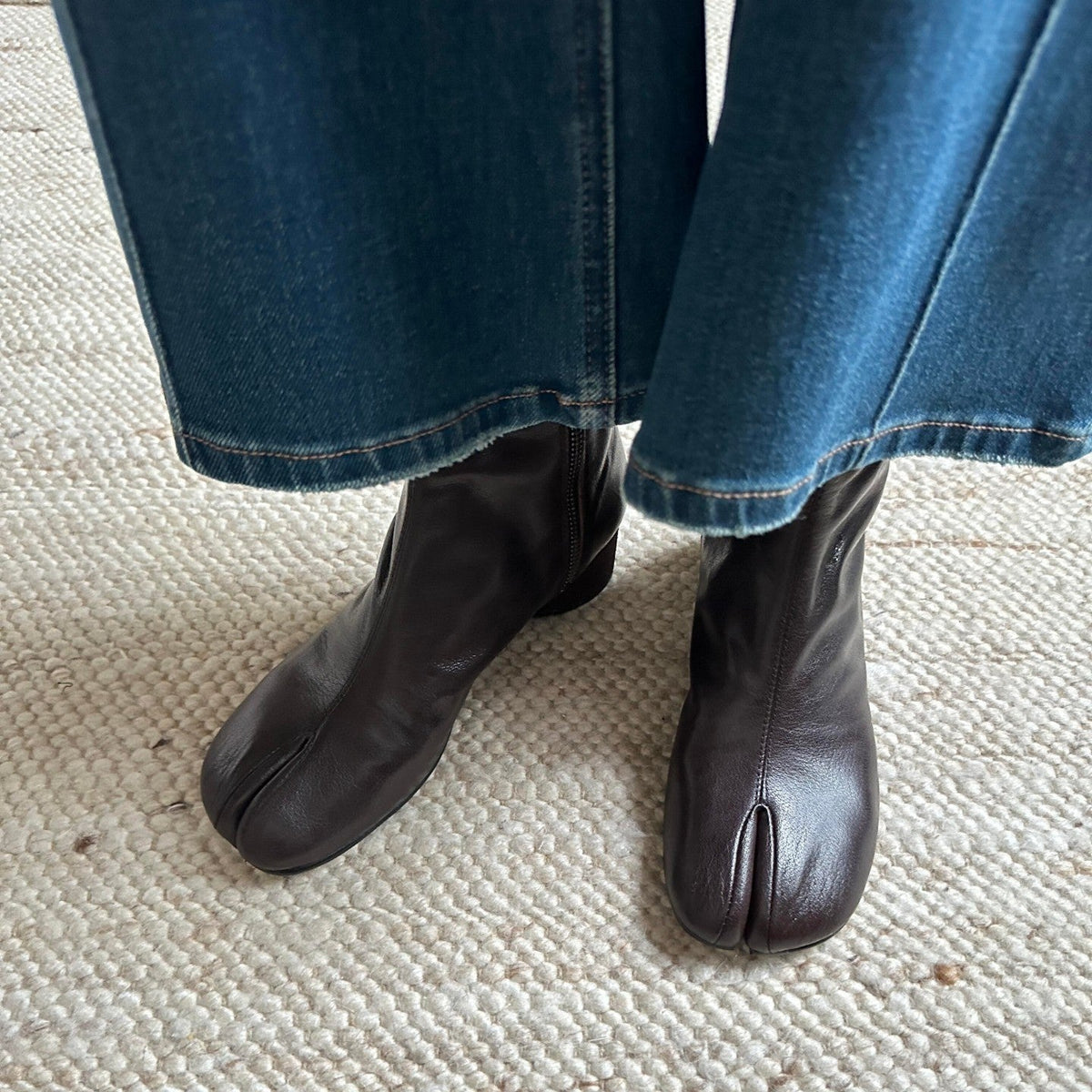 A close-up of a person's feet wearing dark brown leather tabi boots with split toes, paired with blue denim jeans on a textured cream woven rug.