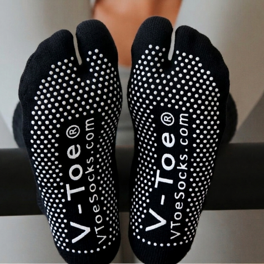 A pair of black V-Toe yoga/pilates Tabi Split-Toe socks with white dotted grip patterns and white branding text, shown on feet resting on a black bar.
