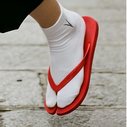 A person walking on a stone street wearing white ankle tabi split toe socks and red-and-white Japanese geta sandals.