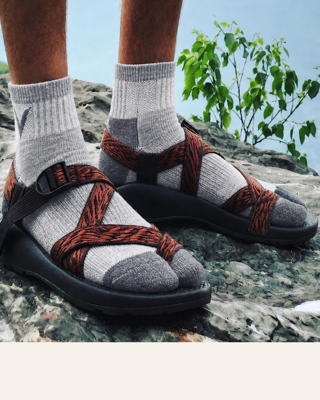 A person wearing gray wool Split-Toe V-TOE socks and colorful sandals with rust-brown woven straps, standing on a rocky outdoor surface with greenery and sky in the background.