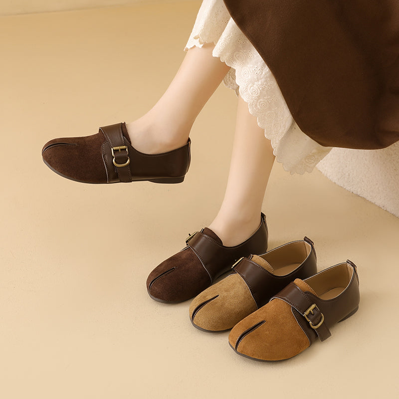 A person wearing brown split-toe loafers with buckle straps, shown with one shoe lifted and two shoes on the floor. The shoes have a suede-like finish in dark brown and tan, with a small gold-tone buckle.