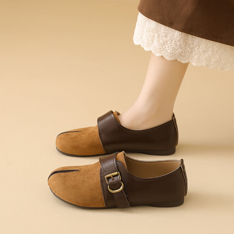 A pair of women's slip-on shoes in brown and tan suede, shown on a model's feet against a warm beige background. The shoes feature a split design with a dark brown leather strap and buckle on one side and a tan suede panel on the other.
