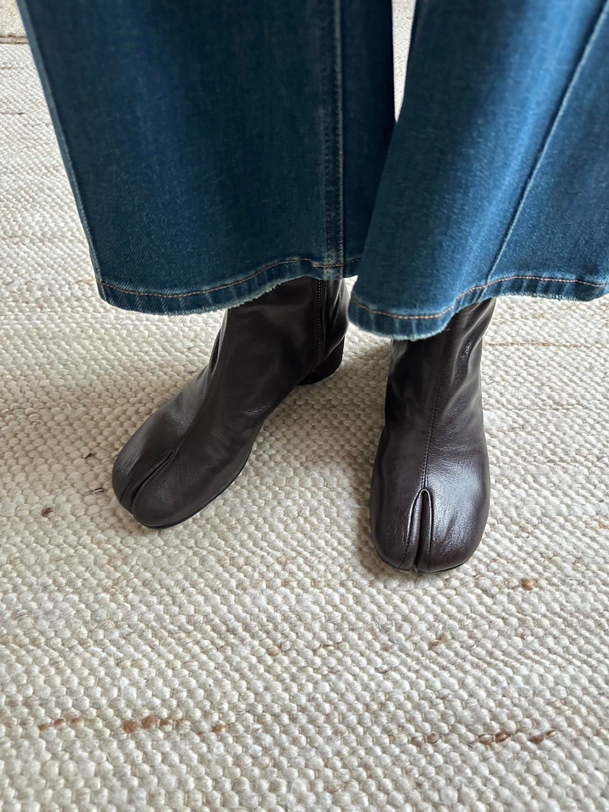 A person wearing dark brown tabi-style leather ankle boots with blue jeans, standing on a textured cream-colored woven rug.