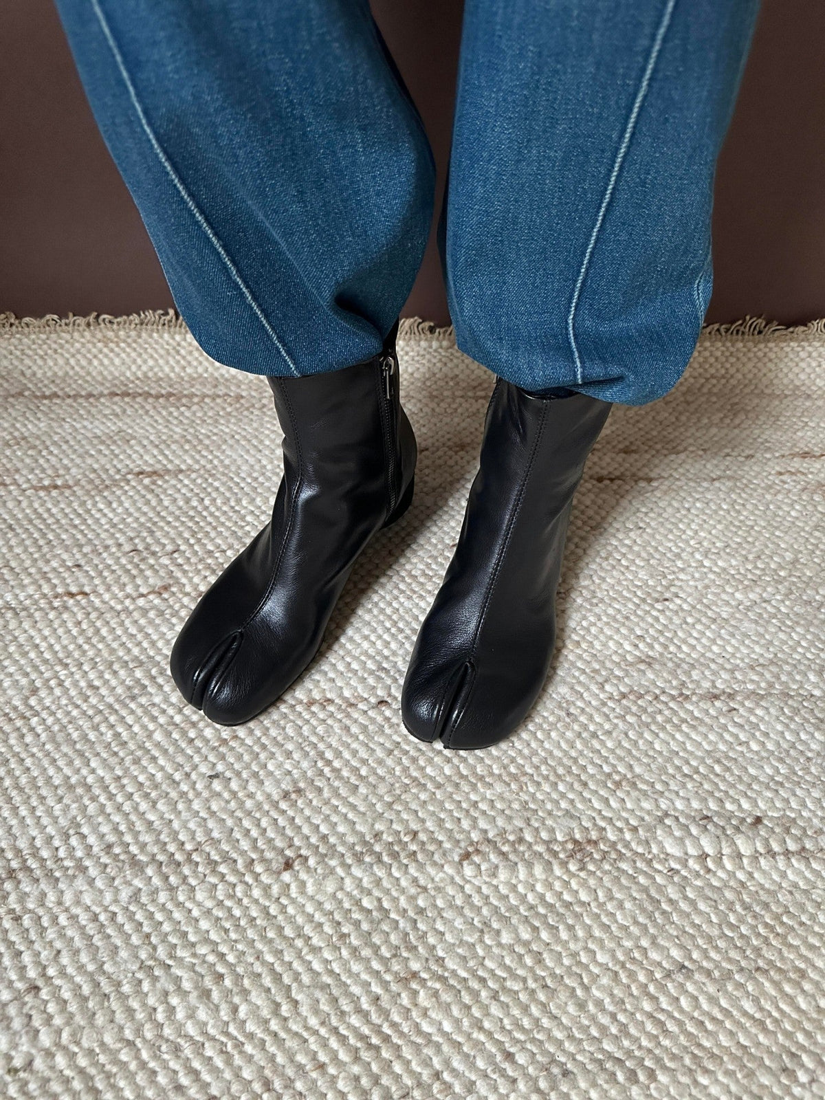 Black leather tabi-style ankle boots worn with blue jeans on a textured beige woven rug.